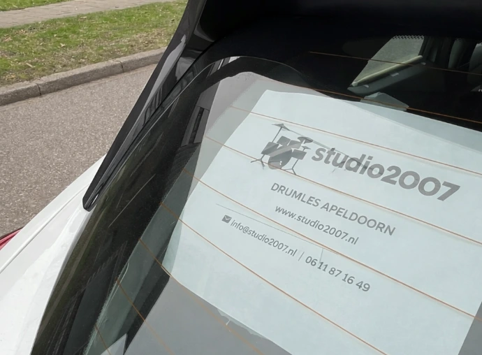 Hier zie je de poster met de tekst 'Studio2007 | Drumles in Apeldoorn |  www.studio2007.nl | info@studio2007.nl' op de achterruit van een witte auto
